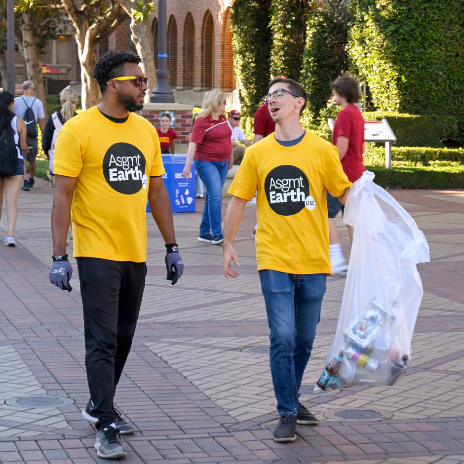 2 USC students wearing Asgmt Earth shirts holding bag of recycled cans on campus